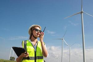 The confident woman in white helmet working with digital tablet and using walkie talkie at a renewable energy farm. Female inspector controlling the functioning of wind turbines outdoors photo