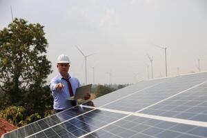 un mayor masculino ingeniero inspecciona solar paneles en el molino granja. limpiar energía. ingenieros inspeccionar un solar panel sistema con un turbina granja en el fondo, un renovable energía concepto foto