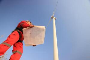 windmill engineer with red safety uniform hold drawing work at front of wind turbine farm photo