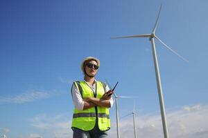 The confident woman in white helmet working with digital tablet and using walkie talkie at a renewable energy farm. Female inspector controlling the functioning of wind turbines outdoors photo