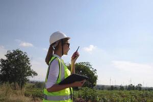 The confident woman in white helmet working with digital tablet and using walkie talkie at a renewable energy farm. Female inspector controlling the functioning of wind turbines outdoors photo