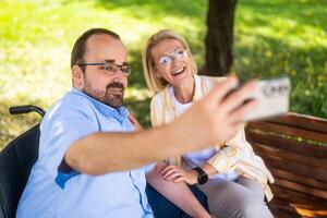 Man in wheelchair is spending time with his mother in park. They are taking selfie. photo