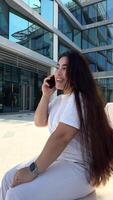 feliz jovem mulher sentado em a Banco em a público área, digitando bate-papo mensagem em Smartphone, sorridente menina usar célula telefone conversando, procurando em formação em navegador sem fio Internet, conectados compras video