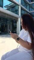 feliz jovem mulher sentado em a Banco em a público área, digitando bate-papo mensagem em Smartphone, sorridente menina usar célula telefone conversando, procurando em formação em navegador sem fio Internet, conectados compras video