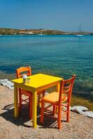 Cafe tableon beach in Adamantas town on Milos island with Aegean sea with boats in background photo