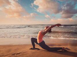 Sporty fit woman practices yoga Anjaneyasana at beach on sunset photo