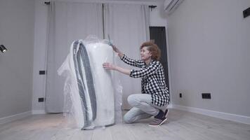 Housewife opens a newly purchased latex mattress from a vacuum roll without air. Mature woman unpacking mattress in new empty apartment without furniture while relocating to new home. Move-in. video