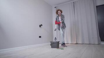 Happy mature housewife using mop and bucket of water, singing and dancing while doing housework in empty room. Cheerful woman enjoying housework, having fun while cleaning apartment before moving in. video