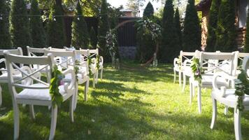 Wedding decor for outdoor cerimony. Wedding arch and chairs for guests in white color on green grass in sunny weather. Backyard wedding venue. Furniture and decoration for outside wedding party. video