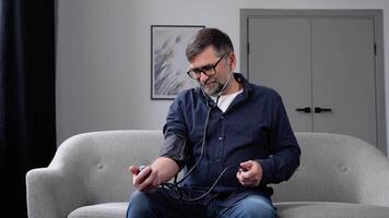 Bearded senior man using modern tonometer at home. A man measuring his blood pressure, making self checkup. Heart problems, heart attack and stroke prevention video