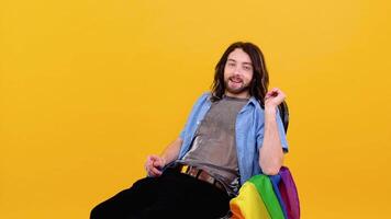 Gay man dressed in mesh t-shirt sits in a chair with a multicolored flag. Concept diversity, transsexual, and freedom video