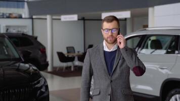 Caucasian man in glasses and suit coming to the camera and speaks by phone in car dealership video