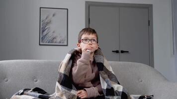 A little boy wrapped in a blanket sprays a nasal spray while sitting on sofa video