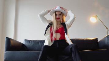 A woman with dyed hair and make-up in a hat, white jacket and red bra poses in the studio. Fashion shooting concept video