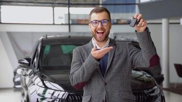 Positive seller smiling for camera and demonstrating keys while standing near new electric car in showroom video