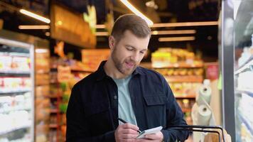 een Mens maakt aantekeningen in de boodschappen doen lijst. jong Mens met trolley vol van producten lezing boodschappen doen lijst video