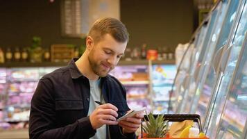 A man makes notes in the shopping list. Young man with trolley full of products reading shopping list video