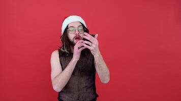 Young sexy gay man in a Santa hat paints lips isolated on red background. People lifestyle fashion lgbtq concept video