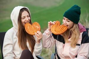 dos joven mujer en calentar ropa teniendo divertido y comiendo galletas en el parque foto