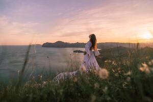 un mujer en un blanco vestir soportes en un herboso colina con vista a el Oceano foto