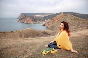 A woman sits on the grass with a bouquet of yellow flowers in her lap. She is looking out at the ocean, taking in the view. Concept of peace and tranquility. photo