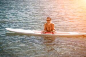 mujer cenar mar viajar. Deportes niña en un tabla de surf en el mar en un soleado verano día. en un negro baños traje, él se sienta en un sapa en el mar. descanso en el mar. foto