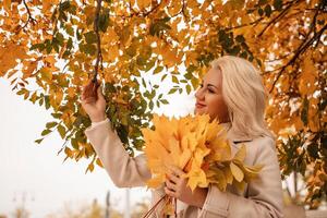 A blonde woman is holding a bunch of yellow leaves in her hand. She is standing in front of a tree with leaves that are changing colors. The scene has a peaceful and serene mood. photo