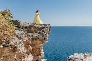 Woman in a yellow dress on the sea. Side view Young beautiful sensual woman in yellow long dress posing on a rock high above the sea at sunset. Girl in nature against the blue sky photo