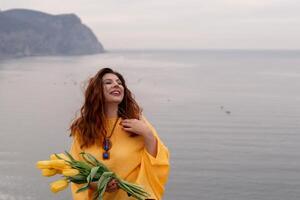 Portrait happy woman woman with long hair against a background of mountains and sea. Holding a bouquet of yellow tulips in her hands, wearing a yellow sweater photo