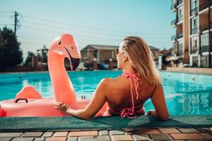 un mujer en un rosado bikini es sentado en un rosado inflable flamenco en un piscina. el escena es juguetón y divertido, con el mujer disfrutando su hora en el agua. foto