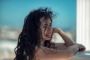 mar mujer descansar. retrato de un mujer con largo Rizado negro pelo en un beige vestir soportes en un balcón en contra el fondo de el mar. turista viaje a el mar. foto