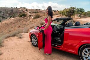 A woman in a red dress stands in front of a red convertible. The scene is set in a desert, with the woman posing for a photo. Scene is playful and fun, as the woman is dressed up. photo