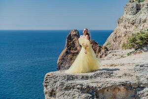 Woman in a yellow dress on the sea. Side view Young beautiful sensual woman in yellow long dress posing on a rock high above the sea at sunset. Girl in nature against the blue sky photo