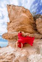 A woman in a red silk dress stands by the ocean, with mountains in the background, as her dress sways in the breeze. photo