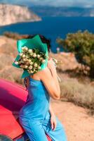 A woman is sitting on a red car with a bouquet of flowers in her hand. The flowers are white and green, and the woman is wearing a blue dress. The scene has a peaceful and serene mood. photo