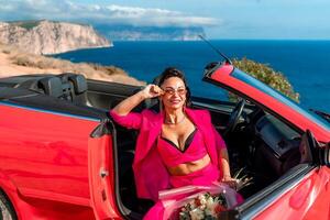 A woman is sitting in the back of a red convertible car. The car is parked on a dirt road near the ocean. The woman is wearing a pink suit and red shoes. Scene is relaxed and carefree. photo