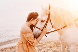 A woman in a dress stands next to a white horse on a beach, with photo