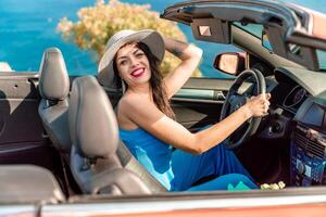 A woman is driving a red convertible with a hat on. She is smiling and she is enjoying the ride. photo