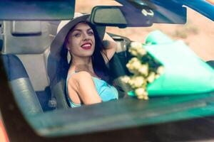 A woman is driving a car with a bouquet of flowers in the back seat. She is wearing a hat and has her eyes closed. photo