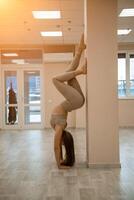 el niña es formación en el yoga estudio. vestido en un beige chandal haciendo un parada de manos en contra el pared. sano estilo de vida y yoga concepto. foto