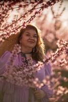 Woman blooming peach orchard. Against the backdrop of a picturesque peach orchard, a woman in a long pink dress and hat enjoys a peaceful walk in the park, surrounded by the beauty of nature. photo