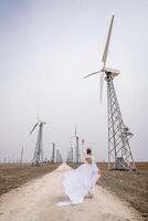 A woman in a white dress is walking down a dirt road in front of a row of wind turbines. photo