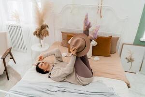 Lifestyle concept. A young middle-aged woman in a sweater lies on the bed and laughs, holds a brown hat in her hands. photo