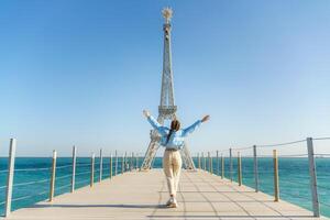 grande modelo de el eiffel torre en el playa. un mujer camina a lo largo el muelle hacia el torre, vistiendo un azul chaqueta y blanco vaqueros. foto