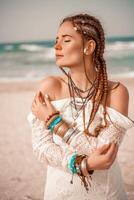 Model in boho style in a white long dress and silver jewelry on the beach. Her hair is braided, and there are many bracelets on her arms. photo