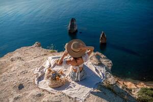 Street photo of a beautiful woman with dark hair in a white top, shorts and a hat having a picnic on a hill overlooking the sea