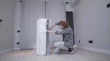 Topic of moving to new apartment. An elderly woman unpacks a new polyethylene orthopedic mattress in an empty apartment. A happy middle-aged female opens the packaging of a new mattress in a new home. video