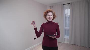 Elderly female real estate agent holds keys to new apartment. Mature woman Realtor property broker gives key to house while standing in background of empty small cozy apartment for rent. Real estate. video