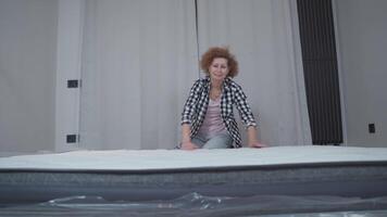 ouderen vrouw verheugd met aankoop nieuw matras in nieuw verhuurd appartement in leeg slaapkamer. volwassen vrouw gelukkig probeert en inspecteert nieuw slapen matras Bij huis enkel en alleen na hebben verhuisd naar nieuw huis. video