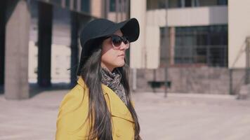 elegante niña en un amarillo Saco con largo oscuro pelo en un sombrero camina abajo el calle toma apagado su lentes y mira adelante. lento movimiento video